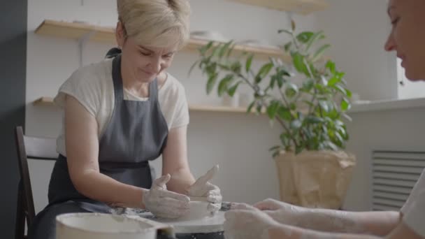 Woman master transfers knowledge to an elderly woman working on a potters wheel and making a mug of ceramics in her workshop in slow motion — Stock Video