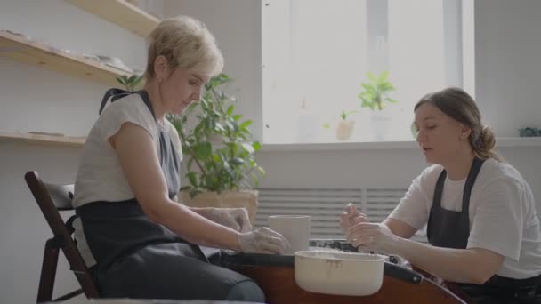Mujer maestra transfiere conocimientos a una anciana que trabaja en una rueda de alfarero y hace una taza de cerámica en su taller en cámara lenta — Vídeo de stock