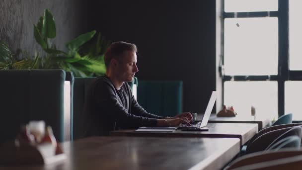 Um homem sério é um estudante de negócios que trabalha estudando sentado em uma mesa e digitando uma mensagem em um laptop. Escrevendo texto no teclado — Vídeo de Stock