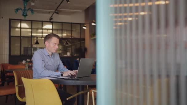 Um homem sério é um estudante de negócios que trabalha estudando sentado em uma mesa e digitando uma mensagem em um laptop. Escrevendo texto no teclado — Vídeo de Stock