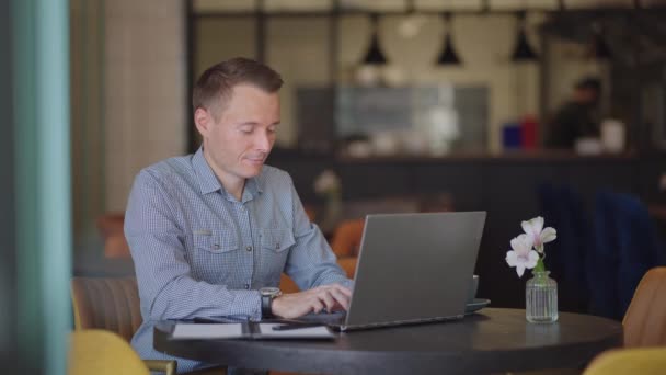 Um homem sério é um estudante de negócios que trabalha estudando sentado em uma mesa e digitando uma mensagem em um laptop. Escrevendo texto no teclado — Vídeo de Stock