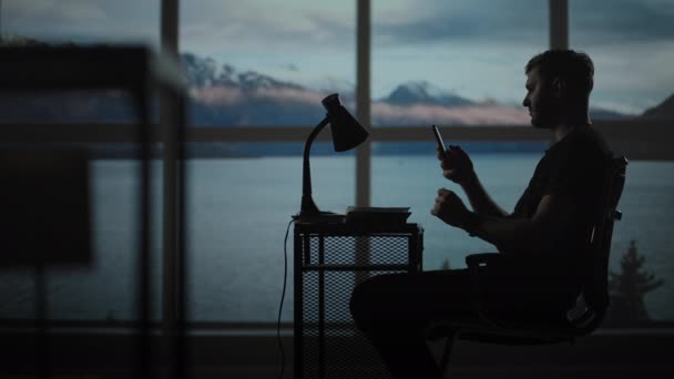 Silhouette d'un homme assis à une table regardant un téléphone portable penser et analyser le marché boursier, résoudre des problèmes personnels, remboursement de prêts, services bancaires sur Internet — Video