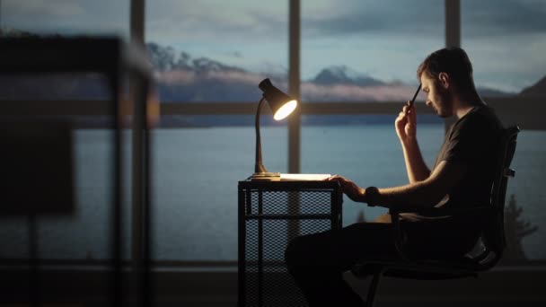 Ciddi şekilde konsantre olmuş genç erkek öğrenci gece lambaların altında rahat işyerinde ders çalışıyor. Genç bir adam düşünür, yaratıcı bir proje yaratır ve bir kalemle yazar.. — Stok video