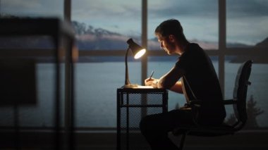 Ciddi şekilde konsantre olmuş genç erkek öğrenci gece lambaların altında rahat işyerinde ders çalışıyor. Genç bir adam düşünür, yaratıcı bir proje yaratır ve bir kalemle yazar.. 