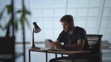 Mobil uygulamalar kullanarak gülümseyen genç adam masada oturuyor. Erkek milenyum profesyoneli ofiste modern akıllı telefon mesajı tutuyor. Genç işadamı iş zamanı için yararlı mobil uygulamalar kullanıyor