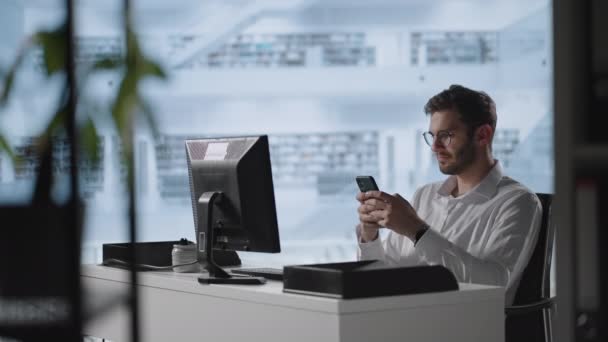 Un jeune homme souriant utilisant des applications mobiles s'assoit à table. Homme millénial professionnel tenant message texte smartphone moderne dans le bureau. Jeune homme d'affaires utilisant des applications mobiles utiles pour le temps d'affaires — Video