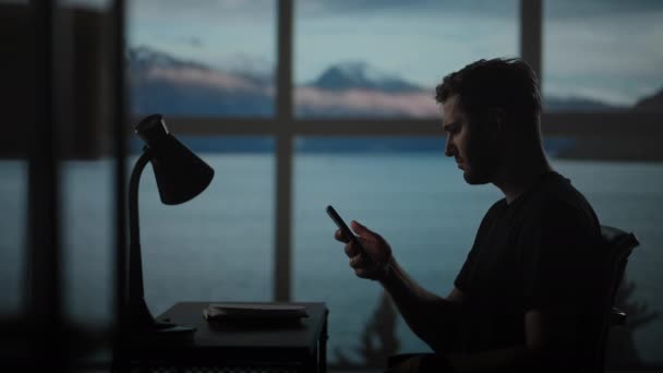 Het silhouet van een bedachtzame man zittend aan een tafel tegen de achtergrond van een raam met oceanen en de zee. Een rusteloze man zittend aan een tafel met een bureaulamp — Stockvideo