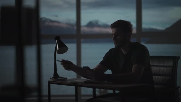 Masasında lambayla oturan düşünceli bir adam kalemle yazar. Gündüz vakti alacakaranlıkta pencerenin arkasında üzgün bir adam düşünür ve yazar. — Stok video