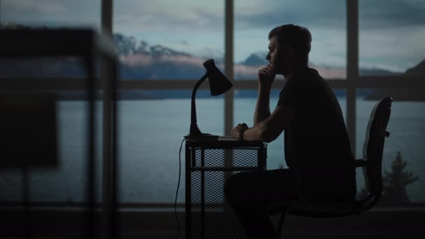 Düşünceli bir adamın silueti, okyanus ve denizin olduğu bir pencerenin arka planında bir masada oturuyor. Masasında lambayla oturan huzursuz bir adam. — Stok video