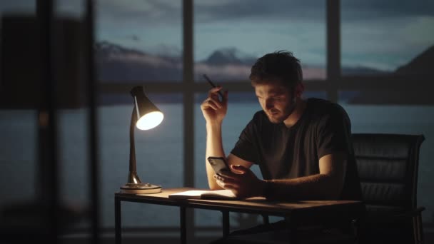 Ein nachdenklicher Mann sitzt an einem Tisch mit Schreibtischlampe und Handy. Der dunkle Tag ist Dämmerung vor dem Hintergrund des Fensters ein trauriger Mann denkt an die Arbeit, während er zu Hause sitzt. — Stockvideo