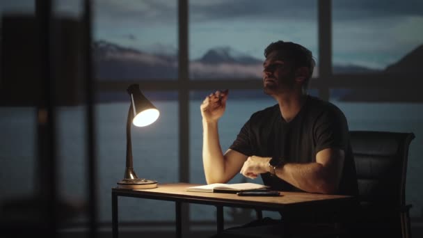 Die Silhouette eines nachdenklichen Mannes, der an einem Tisch vor dem Hintergrund eines Fensters mit Ozeanen und Meer sitzt. Ein unruhiger Mann sitzt an einem Tisch mit einer Schreibtischlampe — Stockvideo