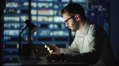 Ofis Binası İş Adamı Cep Telefonu Kullanıyor ve Ofis Penceresinin Yanında Bekliyor. Adam gece geç saatlerde modern ofiste çalışıyor. İşte cep telefonuyla meşgul Kafkas yöneticisi..