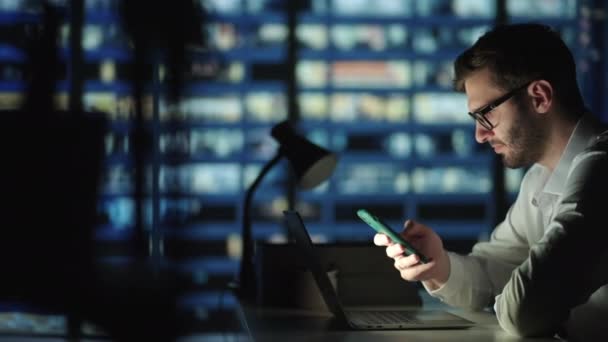 Bürogebäude mit Geschäftsmann, der sein Handy benutzt und am Bürofenster steht. Mann arbeitet spät in der Nacht im modernen Büro. Umtriebiger kaukasischer Manager mit Mobiltelefon am Werk. — Stockvideo