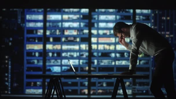 Um jovem cansado a trabalhar num portátil à noite no escritório. Homem de negócios sonolento sentado na mesa no escritório escuro. Empresário cansado e estressado em óculos trabalha em um laptop do escritório da cidade noturna — Vídeo de Stock