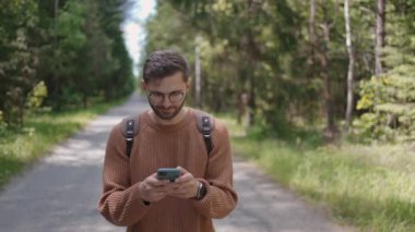 Sırt çantalı genç bir adam ormanda toprak bir yolda yürüyor, mesajlar yazıyor, internette iletişim kuruyor, içeriği görüyor, gülümsüyor..