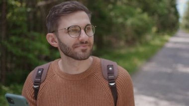 Sırt çantalı genç bir adam ormanda toprak bir yolda yürüyor, araziye bakıyor. Ön görünüm.