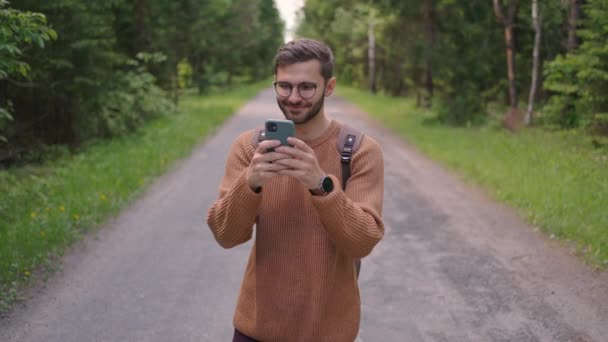 Een man met een rugzak loopt langs een onverharde weg in het bos, schrijft berichten, communiceert online, bekijkt inhoud, glimlacht, verheugt zich. Vooraanzicht. — Stockvideo