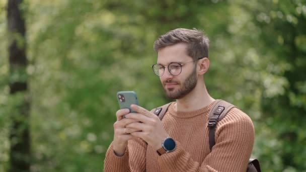 Młody człowiek z plecakiem w lesie, w naturze pisze wiadomości przez telefon komórkowy. Turysta na otwartej przestrzeni komunikuje się w czacie, patrzy na zdjęcia, raduje się, uśmiecha. — Wideo stockowe