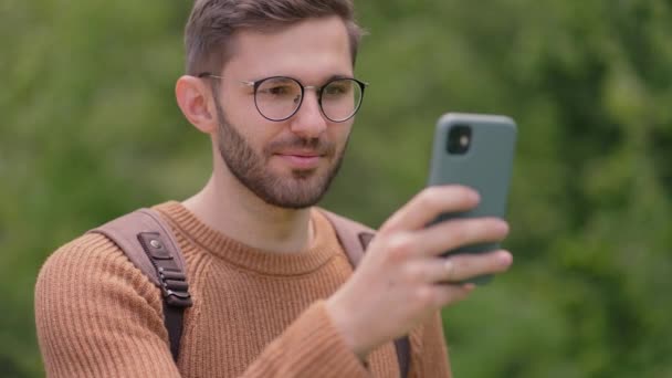 Un hombre con una mochila en una zona forestal en un paseo, en un viaje se comunica a través de un enlace de vídeo en un teléfono móvil. El turista en una videollamada muestra al interlocutor el paisaje, sonríe, se alegra. — Vídeo de stock