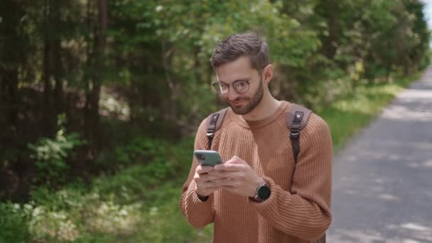 A tourist with a backpack walks along a dirt road in the forest, writes messages, communicates on the Internet, views content. Close-up. — Stock Video