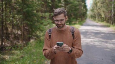 Sırt çantalı genç bir adam orman bölgesinde yolda yürüyor, mesajlar yazıyor, internetten iletişim kuruyor, içeriği görüyor, internette bilgi arıyor, gülümsüyor..