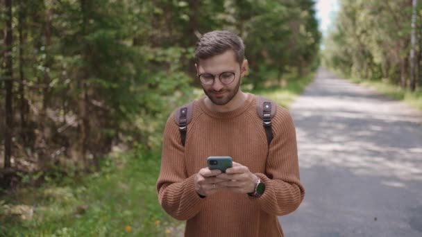 Een jonge man met een rugzak wandelt langs de weg in een bosgebied, schrijft berichten, communiceert op het internet, bekijkt inhoud, zoekt online naar informatie, glimlacht. — Stockvideo