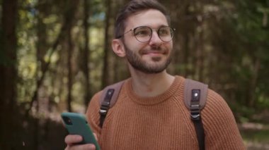 Cep telefonu ve sırt çantası olan bir erkek gezgin ormanda, parkta yürüyor. Bir kişi gülümser, cep telefonunu bilgi aramak için kullanır.