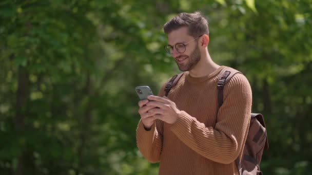 Ένας άνδρας τουρίστας με σακίδιο με κινητό τηλέφωνο σε δασική περιοχή γράφει μηνύματα, επικοινωνεί online, βλέπει περιεχόμενο, χαμογελά, απολαμβάνει μια βόλτα. — Αρχείο Βίντεο