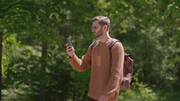 Ein junger Mann mit Rucksack und Handy geht durch den Wald, macht Videos, fotografiert Naturansichten. Erstellung von Videoinhalten, Online-Übertragung. — Stockvideo