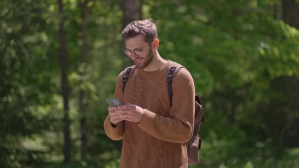 En ung man med en ryggsäck med en mobiltelefon går i skogen, skriver meddelanden, kommunicerar online, tittar på innehåll, ler. — Stockvideo