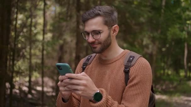 Sırt çantalı ve cep telefonu olan genç bir adam orman bölgesinde bir yol boyunca yürüyor, mesajlar yazıyor, internetten iletişim kuruyor, içeriği görüyor, gülümsüyor, yürümekten hoşlanıyor.. — Stok video