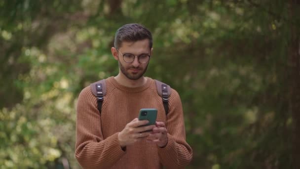 Een mannelijke reiziger met een rugzak en een mobiele telefoon loopt door het bos en probeert een signaal op te vangen. Zoek naar een signaal op een mobiele telefoon in een bosgebied. — Stockvideo