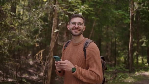 En ung man med en ryggsäck med mobiltelefon går längs en väg i ett skogsområde, skriver meddelanden, kommunicerar online, tittar på innehåll, ler, njuter av en promenad, ser sig omkring. — Stockvideo