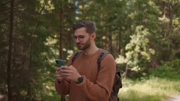 Un viajero masculino con una mochila camina a lo largo de un camino de tierra en el bosque, escribe mensajes, se comunica en Internet, ve contenido. — Vídeos de Stock