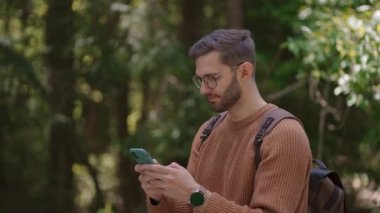 Sırt çantalı bir erkek gezgin ormanda toprak bir yolda yürüyor, mesajlar yazıyor, internetten iletişim kuruyor, içeriğini görüyor, telefonuna notlar alıyor, etrafa bakıyor..