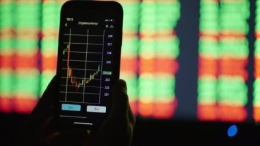 Elinde cep telefonu olan eller, borsa verilerini kontrol ediyor. Cep telefonu borsası. Arkaplanda grafikler ve tablolar