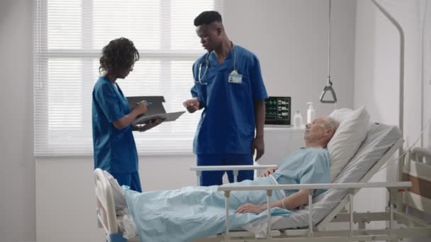 Dua dokter Afrika-Amerika seorang pria dan seorang wanita memeriksa dan berbicara dengan seorang pasien tua yang terbaring di tempat tidur di rumah sakit — Stok Video