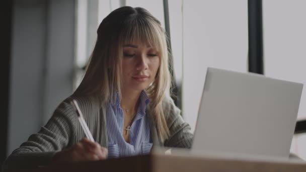 Mujer asiática trabajando en su portátil y escribiendo en su cuaderno, sentada en una mesa. Trabajando en una cafetería. mujer mirando a la pantalla de un ordenador portátil y tomando notas en su cuaderno. estudiar en línea. — Vídeo de stock