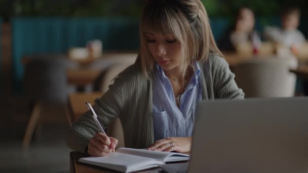 Asyalı bir kadın dizüstü bilgisayarıyla çalışıyor ve onun defterine yazı yazıyor, masada oturuyor. Kahve dükkanında çalışıyorum. Kadın dizüstü bilgisayar ekranına bakıyor ve not defterine not alıyor. çevrimiçi çalışma. — Stok video