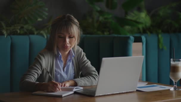 Mulher asiática trabalhando em seu laptop e escrevendo em seu caderno, sentado a uma mesa. A trabalhar num café. mulher olhando para uma tela de laptop e fazendo anotações em seu caderno. estudar online. — Vídeo de Stock