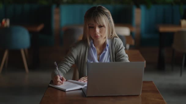 Азиатка работает над своим ноутбуком и пишет в его блокноте, сидит за столом. Работает в кофейне. женщина смотрит на экран ноутбука и делает заметки в блокноте. изучение онлайн. — стоковое видео