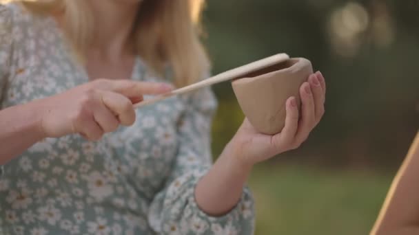 Une jeune femme sculpte intensément un objet en argile dans un espace ouvert, dans la nature, profitant du processus de gros plan. La fille tape le produit d'argile avec une spatule, un outil. — Video