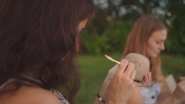 Mladé ženy aplikují vzor, kresbu na hliněné výrobky s pomocí nástrojů, hole na louce, v přírodě, v otevřeném prostoru. Žena zdobí výrobek v close-up. — Stock video