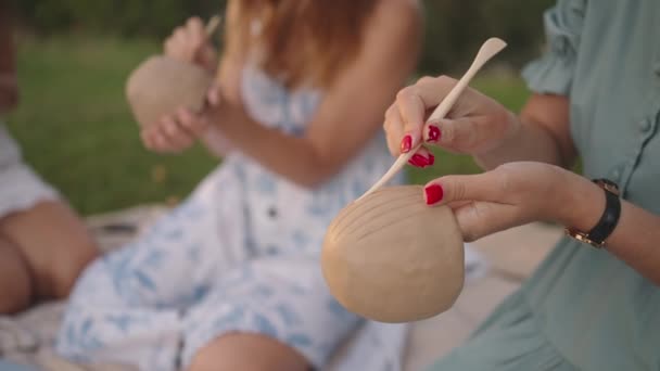 Un grupo de mujeres jóvenes se está concentrando en aplicar patrones a los productos de arcilla con la ayuda de herramientas en un prado, en la naturaleza, en un espacio abierto. Las manos de las mujeres decoran el producto en primer plano. — Vídeo de stock