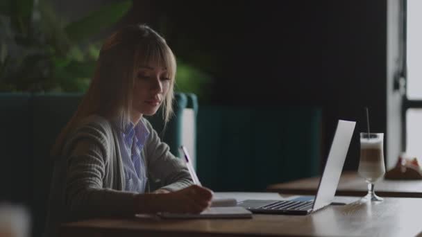 Una studentessa asiatica scrive una lezione online nel campus con una penna. Seduto in biblioteca, una studentessa coreana ascolta una lezione — Video Stock