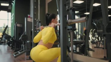 Spor salonunda ya da fitness kulübünde Inflight Fitness Smith Machine kullanarak vücut kasları geliştiren İspanyol genç kadın. Kız omuzlarında halterle ağırlık kaldırırken, çömelmeler sağlık için egzersiz yapıyor..