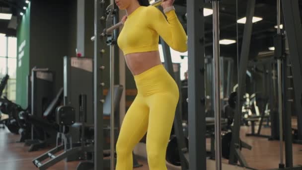 在健身房或健身俱乐部使用Inflight Fitness Smith Machine锻炼西班牙裔年轻女性的肌肉。女子举重时肩膀上戴着杠铃，而蹲姿运动则有利于健康. — 图库视频影像