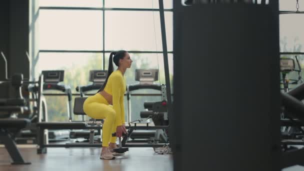 Junge hispanische Brünette hockt in einem gelben Trainingsanzug mit einer Hantel in der Turnhalle — Stockvideo