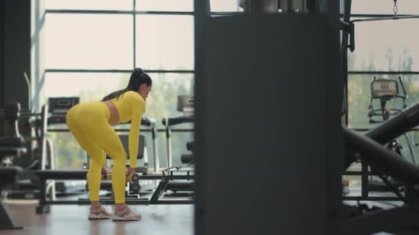 Fit ισπανόφωνη γυναίκα που εκτελεί άρση βαρών deadlift άσκηση με αλτήρα στο γυμναστήριο σε κίτρινο αθλητικό ένδυμα. γυναίκα μελαχρινή γυμναστήριο εκτέλεση κάνει deadlift άσκηση με dumbbell — Αρχείο Βίντεο
