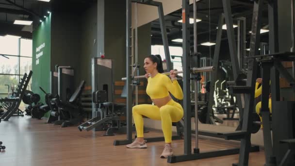 Spor salonunda ya da fitness kulübünde Inflight Fitness Smith Machine kullanarak vücut kasları geliştiren İspanyol genç kadın. Kız omuzlarında halterle ağırlık kaldırırken, çömelmeler sağlık için egzersiz yapıyor.. — Stok video